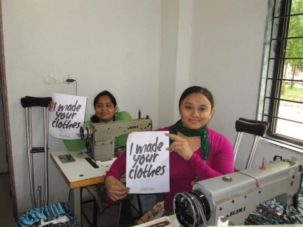 Two women sitting behind sewing machines, holding up signs that read "I made your clothes."